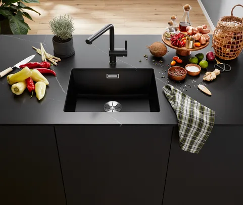 Modern kitchen counter with black sink, colorful peppers, spices, a knife, a plant, and a checkered cloth.