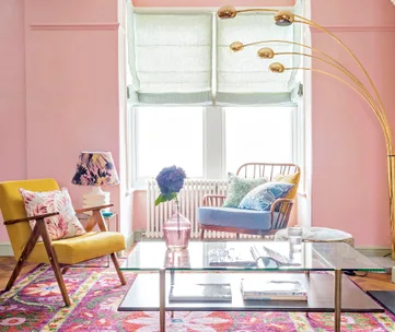 Bright living room with pink walls, colorful furnishings, floral rug, and large arched lamp; two armchairs by window.