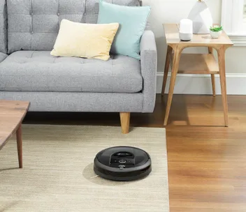 A black Roomba vacuum cleaning a beige rug in a modern living room with a grey sofa and light wood furniture.