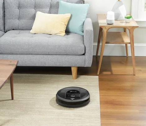 A black Roomba vacuum cleaning a beige rug in a modern living room with a grey sofa and light wood furniture.