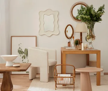 Modern room with wooden table, beige chair, mirrors, artwork, and greenery in a vase. Minimalist decor and neutral tones.