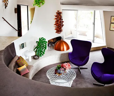 Modern living room with curved sofa, two purple chairs, copper pendant lamp, and glass table with flowers.