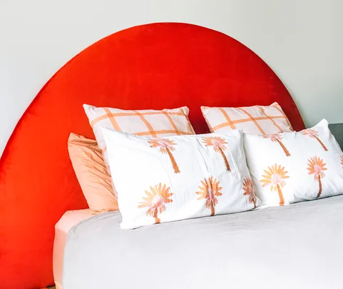 Red semicircular headboard with white palm tree pillows and beige checks pillows on a made bed.