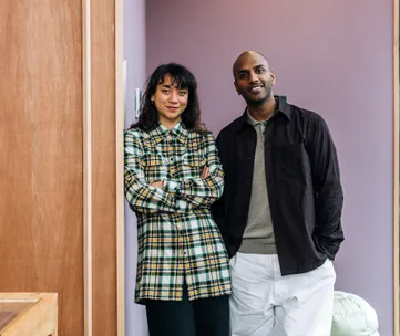 Two people smiling and standing side by side against a purple wall; one in a plaid shirt, the other in dark clothing.