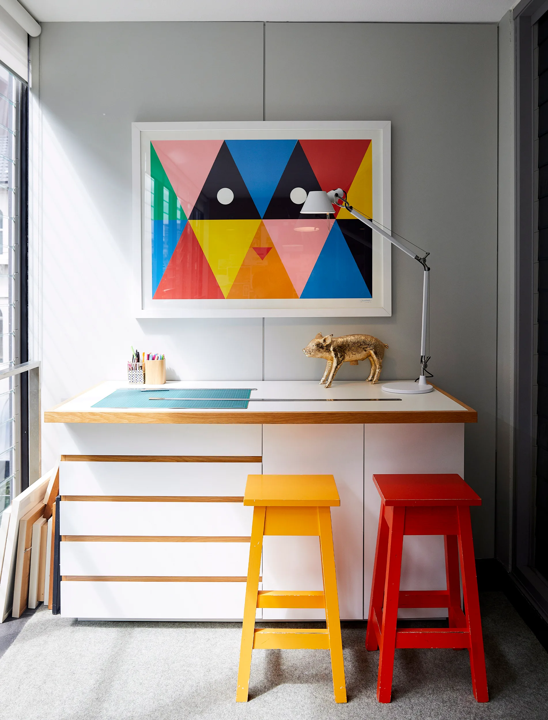 Workspace with colorful geometric artwork, yellow and red stools, desk lamp, and a decorative gold animal statue.