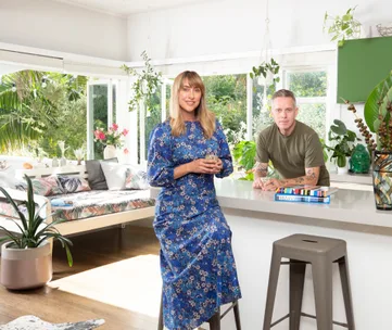 A woman in a blue floral dress and a man in a green shirt in a bright, plant-filled living room.