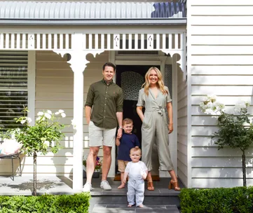 A family of four stands on the porch of a white house with decorative trim and roses.