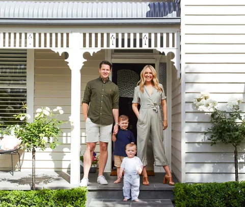 A family of four stands on the porch of a white house with decorative trim and roses.