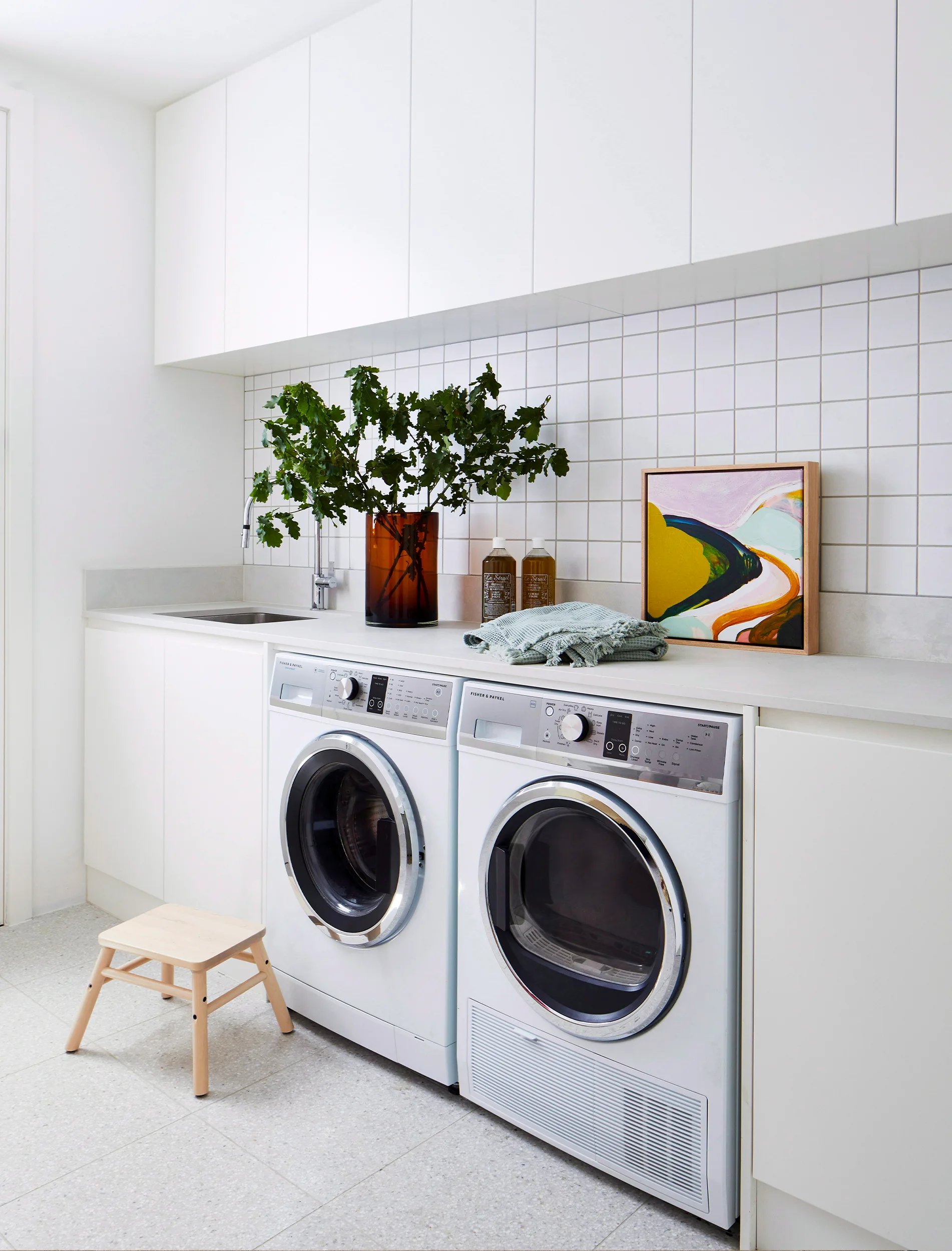 Laundry room with white cabinets, front-load washer and dryer, plant in brown vase, and abstract painting on counter.
