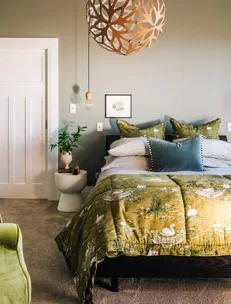 Cozy bedroom with green swan-patterned bedding, modern wooden pendant light, potted plant, and neutral decor.