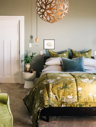 Cozy bedroom with green swan-patterned bedding, modern wooden pendant light, potted plant, and neutral decor.