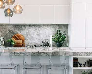 Modern kitchen with marble countertops, pendant lights, transparent chairs, plants, and kitchen appliances.
