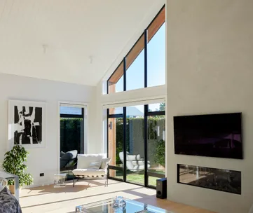Modern living room with large windows, a TV above a fireplace, abstract art, and a chair near a potted plant.