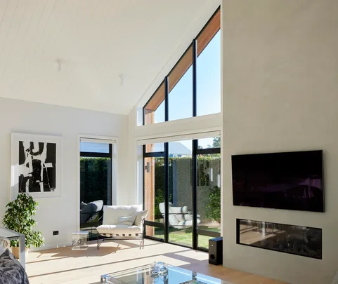 Modern living room with large windows, a TV above a fireplace, abstract art, and a chair near a potted plant.