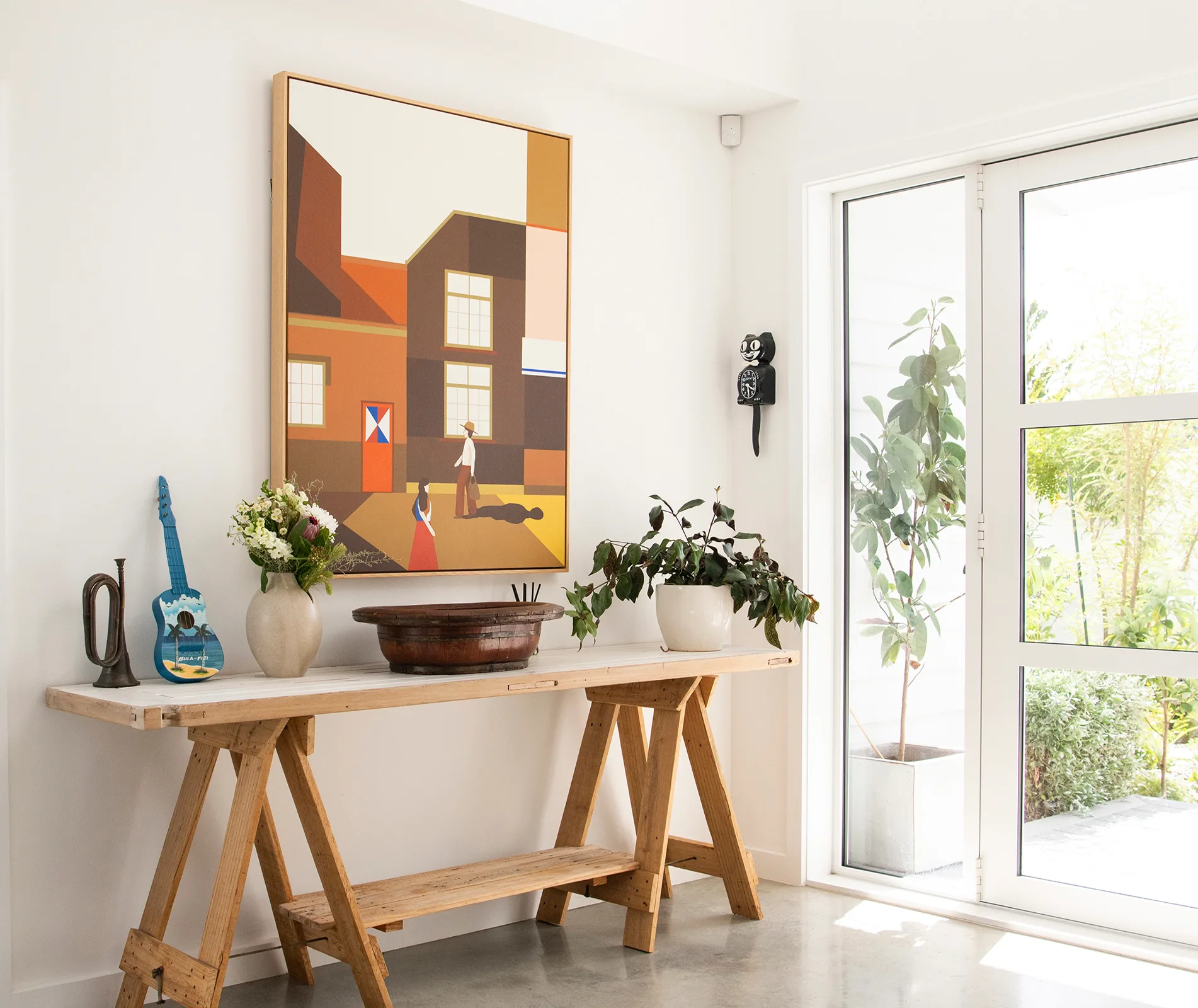 Interior with a painting, wooden table with decor items, plants, and natural light streaming through a window.