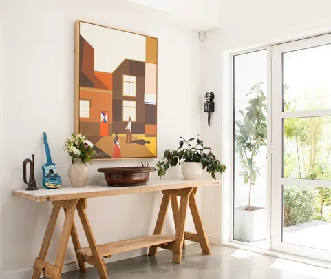 Interior with a painting, wooden table with decor items, plants, and natural light streaming through a window.