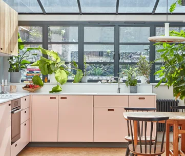 Bright kitchen with skylight, pink cabinets, houseplants on windowsill, books, and wooden chairs around a table.