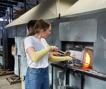 Person in a workshop using metal tools to work with glass near a furnace.