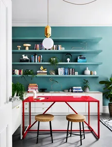 Modern home office with a vibrant red desk, two stools, bookshelves, and a gold-accented pendant light.