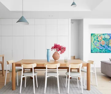 Modern dining room with wooden table, white chairs, a blue vase, wicker vase with red flowers, and colorful wall art.