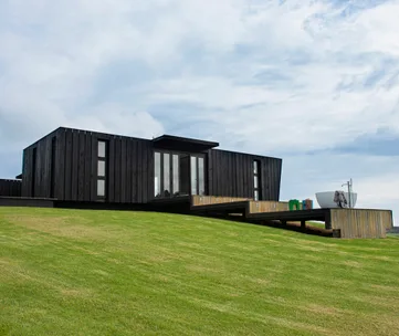 Black modern house with sloped lawn and outdoor tub under a cloudy sky.