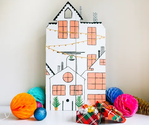 Cardboard house painting with windows, string lights, and presents. Colorful tissue paper balls and ornaments surround it.