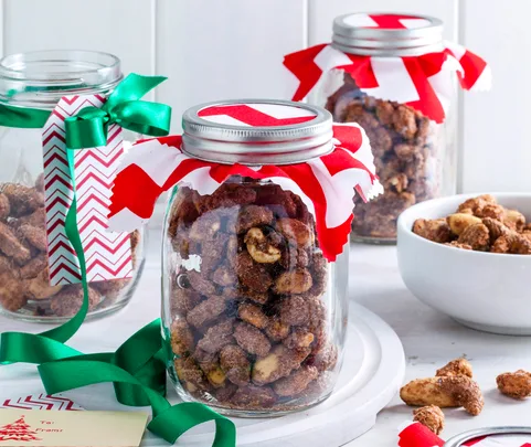 Jars of spiced nuts with festive red and green ribbons and tags, set on a white surface with a small bowl of nuts.