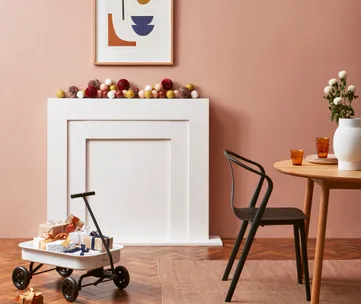 Minimalist room with a white mantel, colorful decor, table, chair, and a toy cart with wrapped gifts on wood flooring.