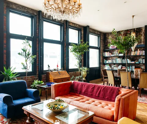 Living room with large windows, vintage furniture, lush plants, and a bookshelf.