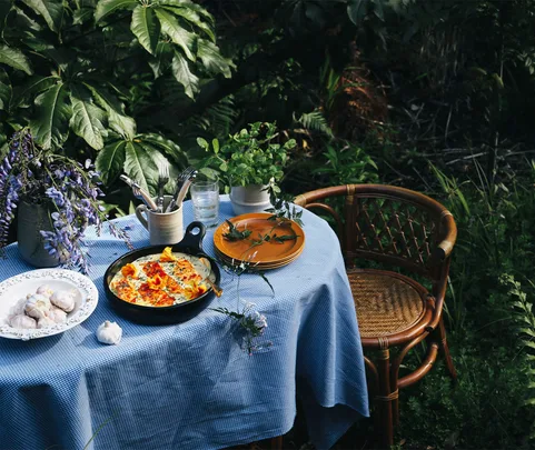 Outdoor table with blue cloth, frittata in pan, plates, utensils, flowers, and herbs. Rattan chair beside.