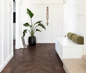 Modern hallway with herringbone wood floor, potted plant, white walls, a cross with rosary, and bench with green cushion.