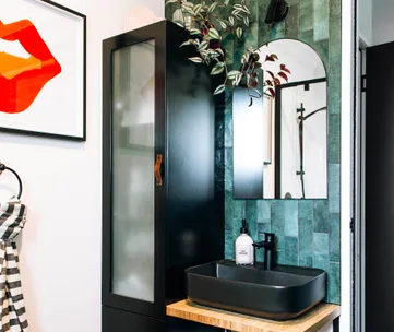 Bathroom with black sink, green tile, round mirror, black cabinet, framed orange lips art, and striped towel.