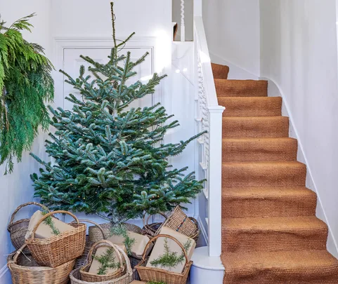 Alt text: "Christmas tree beside staircase with wicker baskets and greenery in a bright hallway."