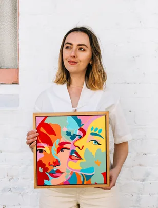Woman holding a colorful abstract painting of faces against a white brick wall.