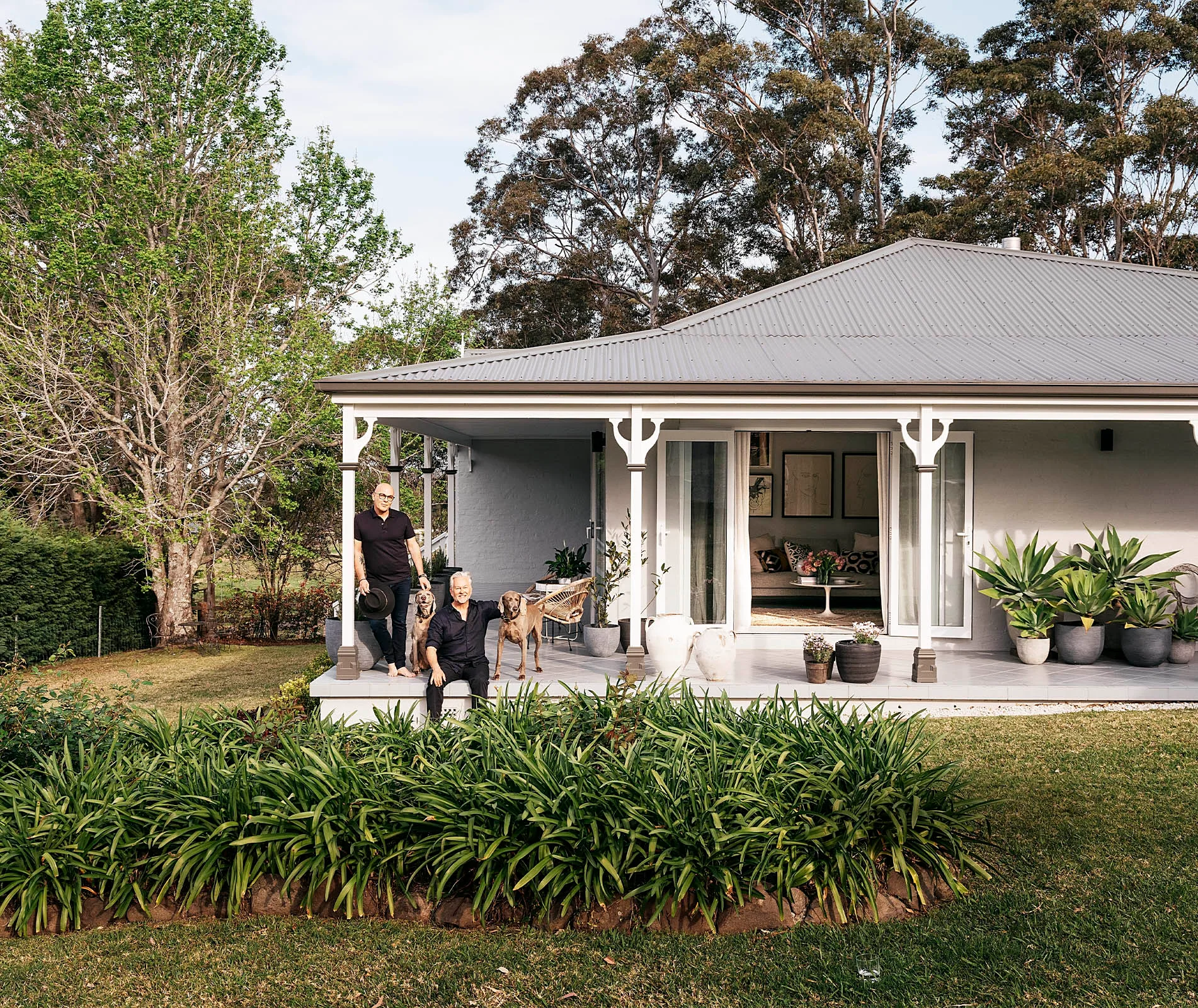 Two people with dogs on porch of a cottage, surrounded by plants and trees, with a glimpse into a cozy interior.