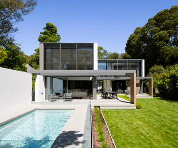 Modern two-story house with glass walls, a patio, and a rectangular pool, surrounded by lush greenery under a clear sky.
