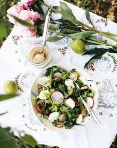 Salad with radishes, olives, mozzarella on embroidered cloth, with pink flowers and lime beside a jar of dressing.