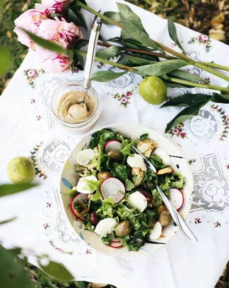 Salad with radishes, olives, mozzarella on embroidered cloth, with pink flowers and lime beside a jar of dressing.