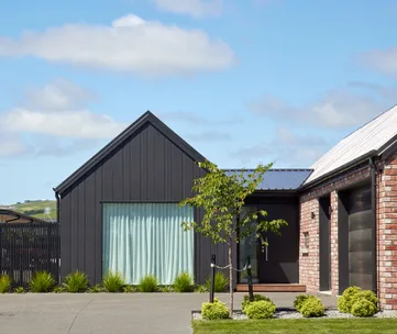 Gabled black and brick modern house exterior with a blue sky and small tree in the foreground.