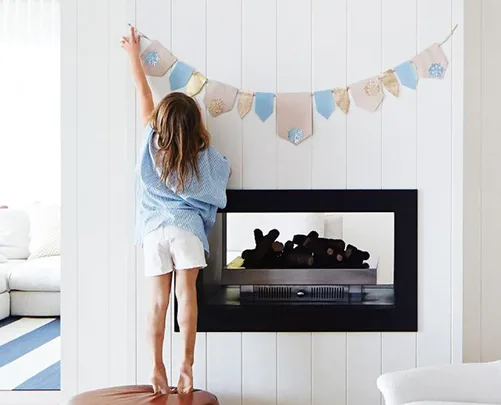 A child in a blue shirt stands on a stool, reaching to hang a colorful banner above a modern fireplace.