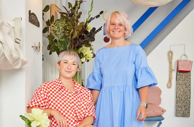 Two women smiling in a bright room with floral and modern decor; one seated, wearing red checkered, the other in blue.
