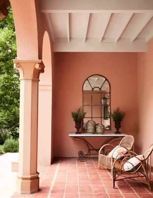 Covered porch with terracotta tiles, wicker chairs, a table with vases, plants, and a mirror on a peach-colored wall.