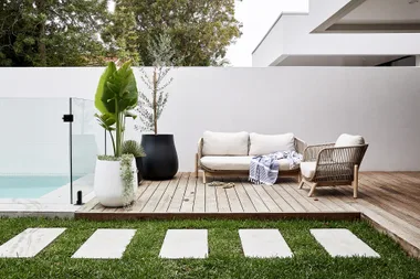 Modern outdoor patio with white sofa, glass pool fence, potted plants, and grassy path.