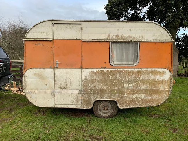 Old, weathered caravan with orange and white paint, parked on grassy area near a tree, next to a Toyota vehicle.