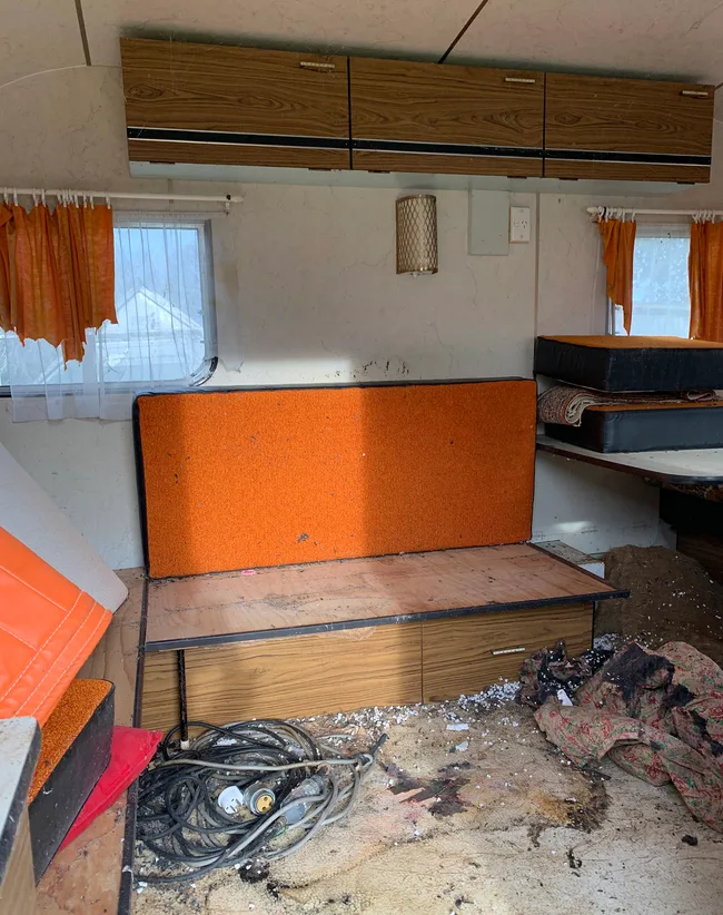 Interior of an old, messy RV with orange seating, tangled electrical cords on a dirty floor, and orange curtains.