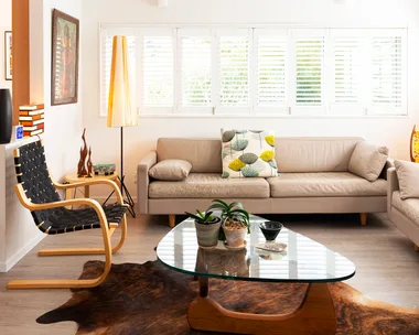Contemporary living room with beige sofas, a glass coffee table, indoor plants, and a cowhide rug under natural light.