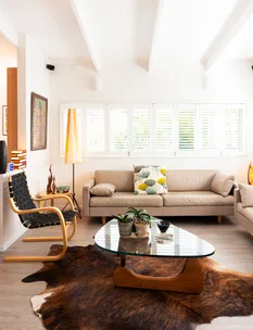 Contemporary living room with beige sofas, a glass coffee table, indoor plants, and a cowhide rug under natural light.