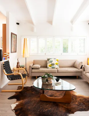 Contemporary living room with beige sofas, a glass coffee table, indoor plants, and a cowhide rug under natural light.