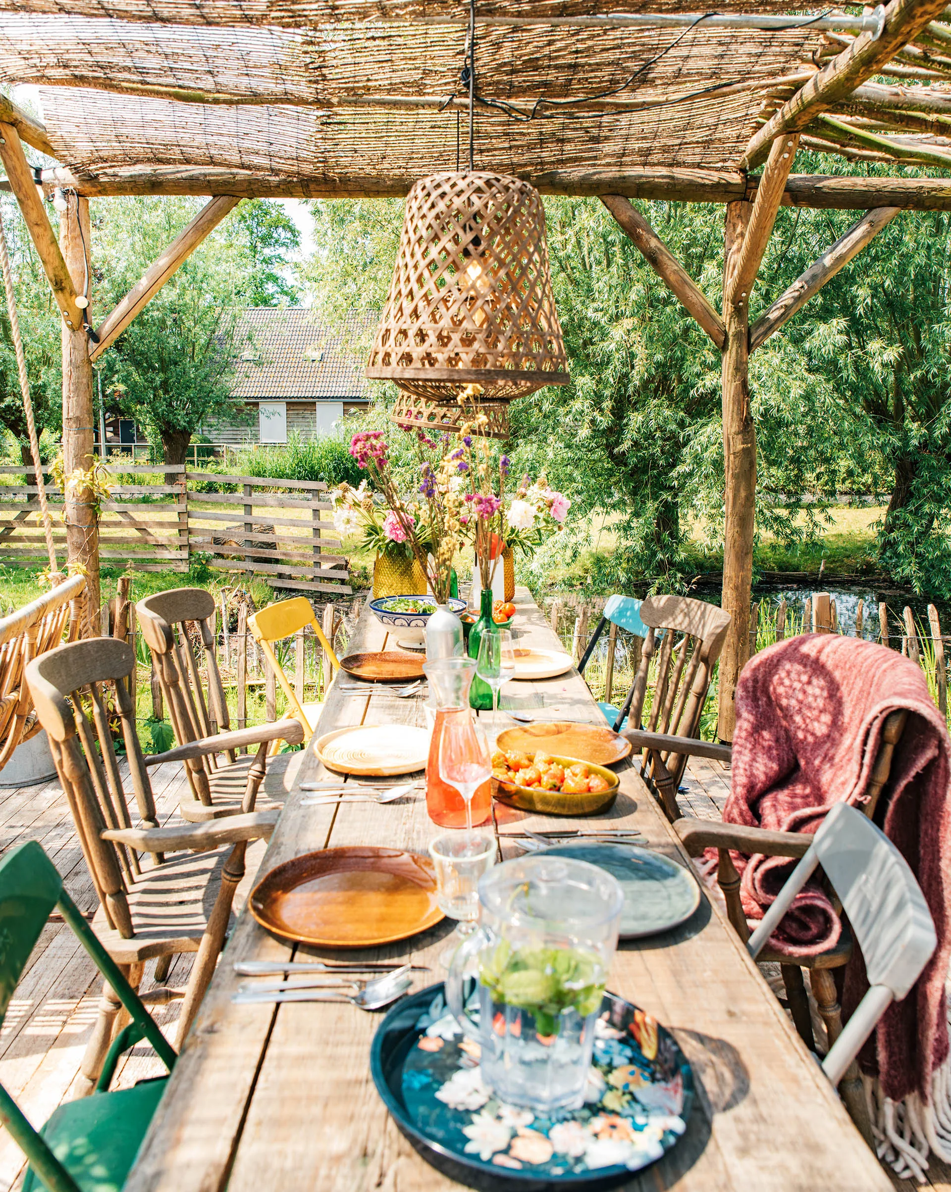 Outdoor wooden dining table under pergola, set for a meal with colorful plates, glasses, and flower vase in rustic garden.