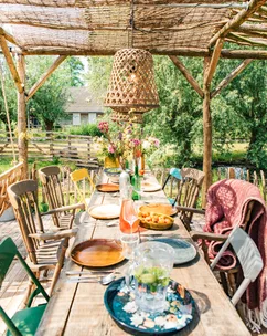Outdoor wooden dining table under pergola, set for a meal with colorful plates, glasses, and flower vase in rustic garden.